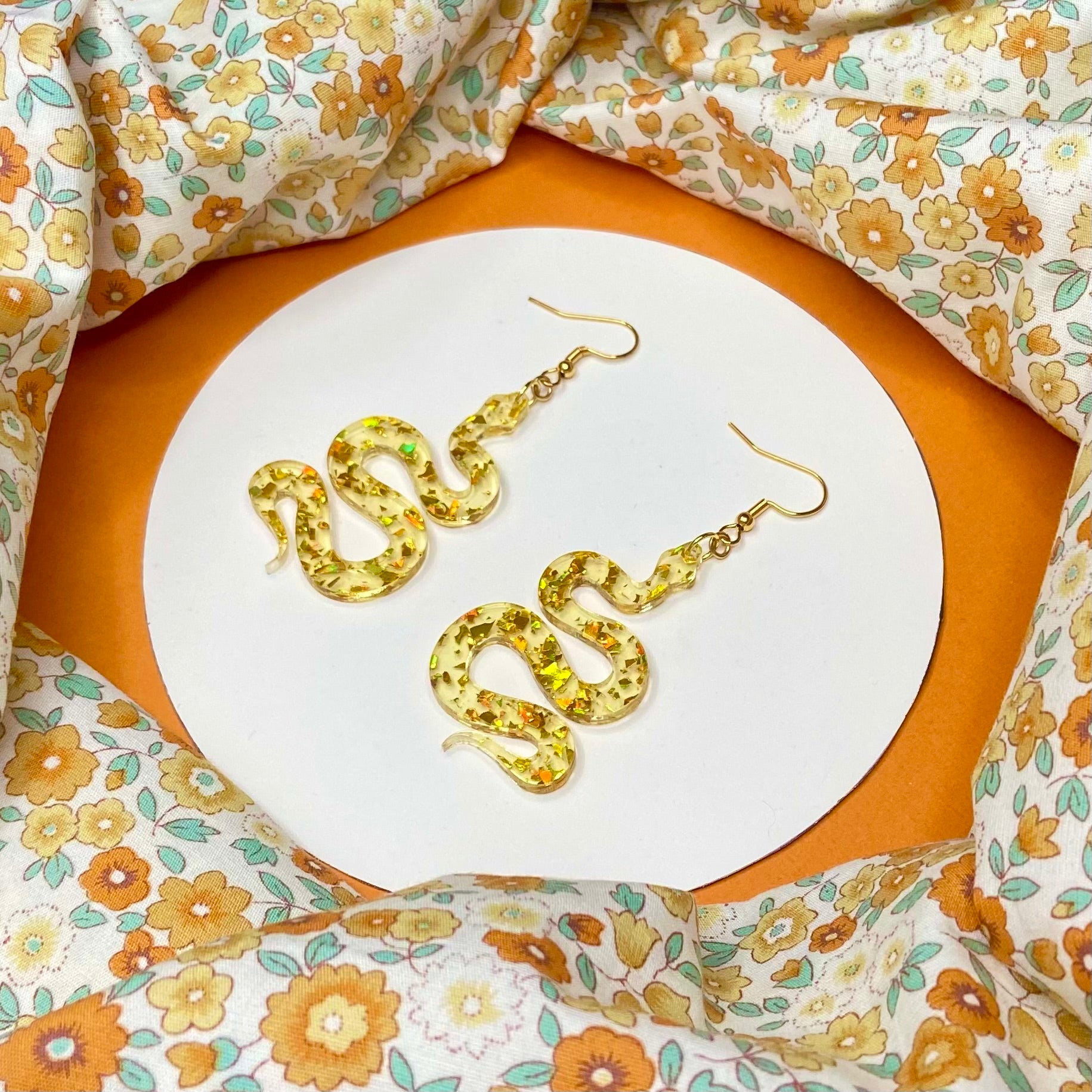 slithering snake shaped acrylic hung from a golden ear wire. The acrylic is clear with flakes of reflective gold throughout it.