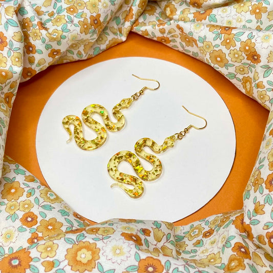 slithering snake shaped acrylic hung from a golden ear wire. The acrylic is clear with flakes of reflective gold throughout it.