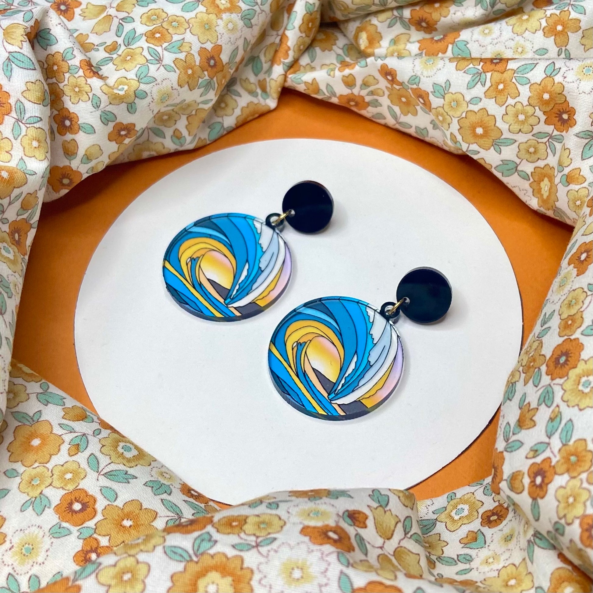 Circular earrings that mimic stained glass. The picture is the tunnel of a wave and through it you can see the sun set over a mountain. It has black circular stud tops or is available as an ear wire/hook.