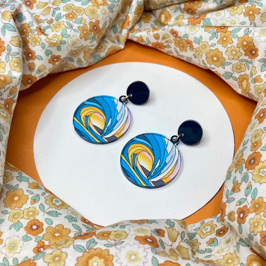 Circular earrings that mimic stained glass. The picture is the tunnel of a wave and through it you can see the sun set over a mountain. It has black circular stud tops or is available as an ear wire/hook.